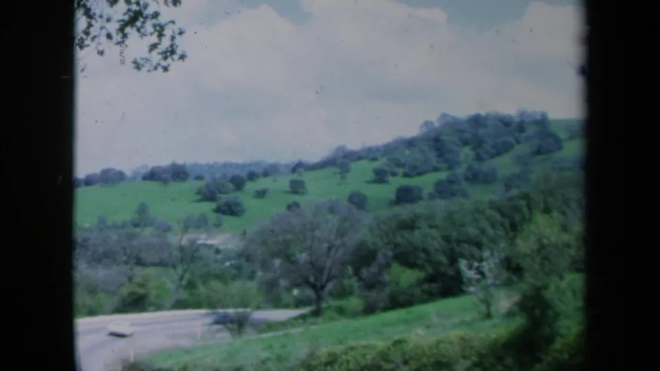 Soft 1969 California 8mm frame of rolling green hills, scattered trees, and a partly visible road below.