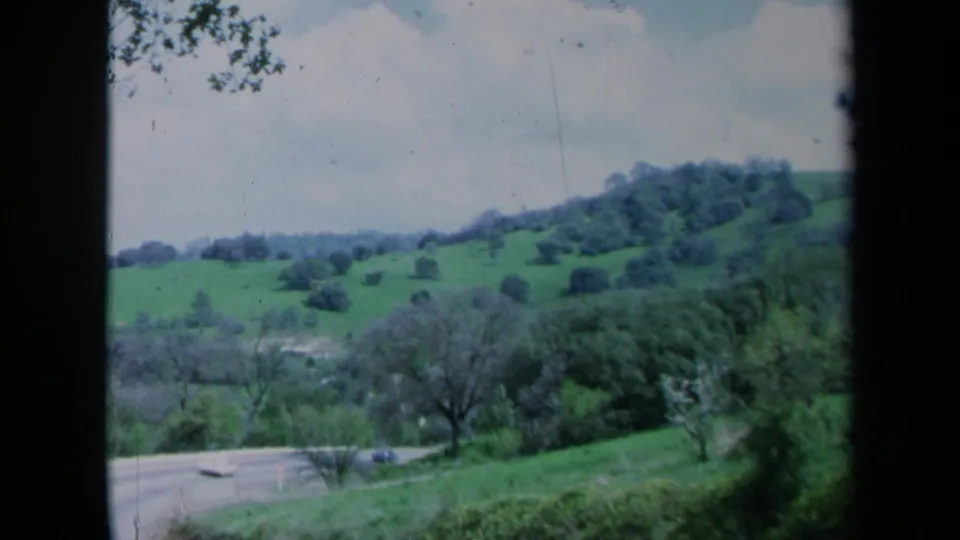 1969 California home-movie view of a green hillside and scattered trees above a curving road with a car.