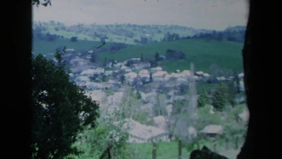 1969 California home-movie frame showing a distant rural town among green hills, partly framed by dark trees and shrubs.