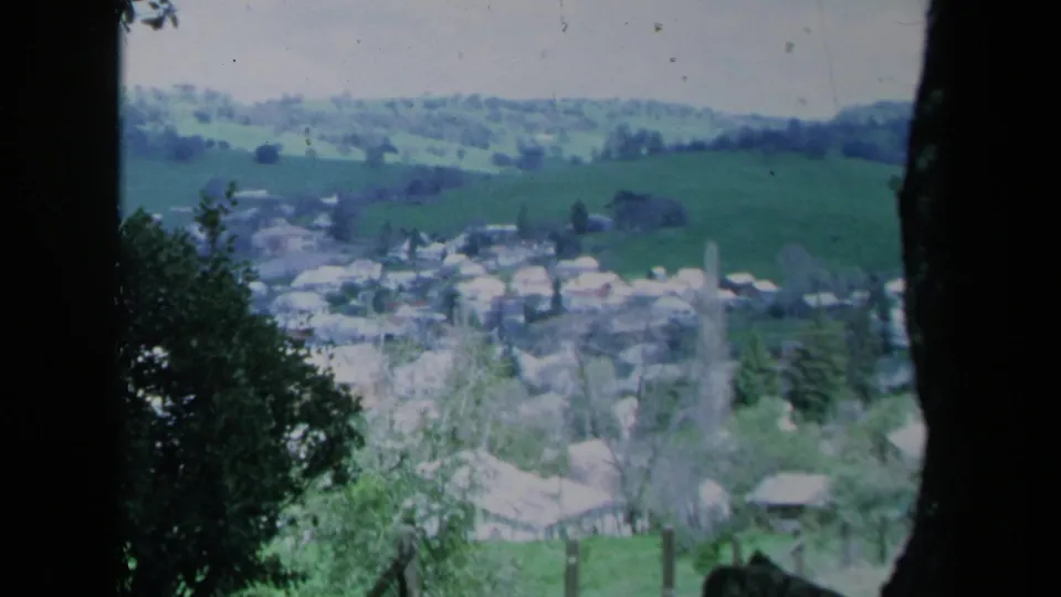 1969 California 8mm view of a rural hillside town, with pale rooftops, fields, and dark foreground foliage.