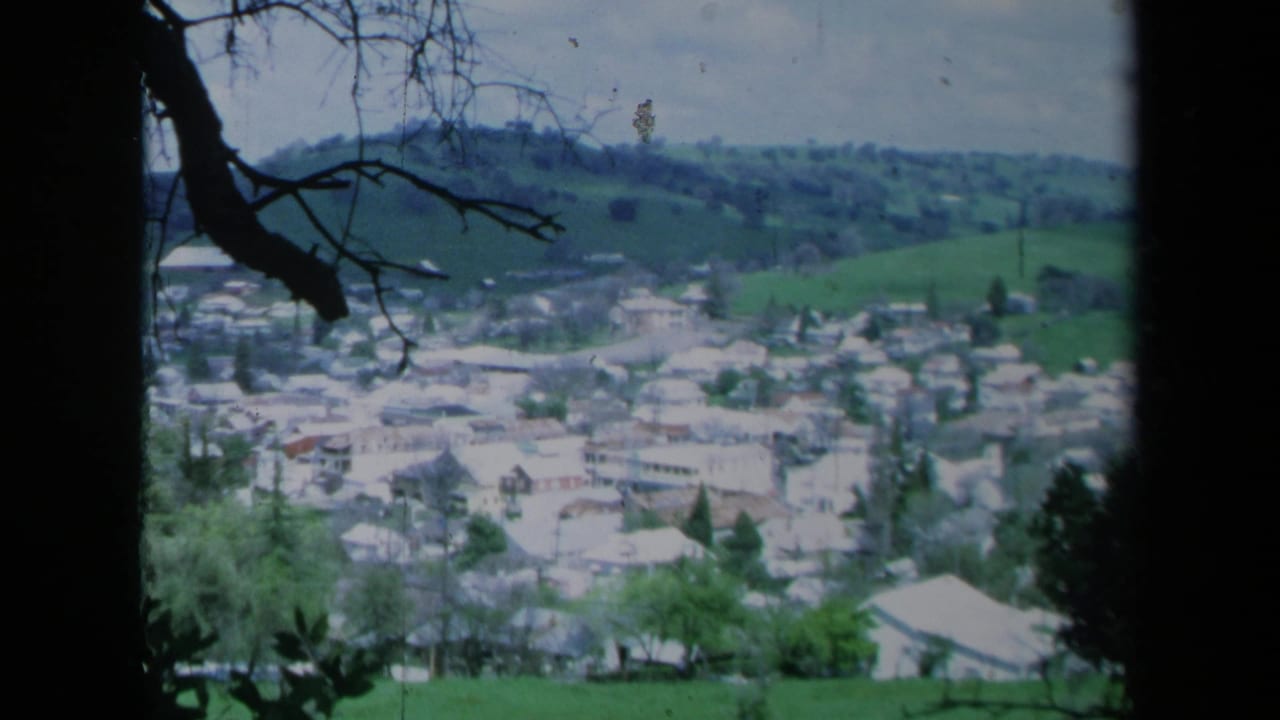 1969 California home-movie view of a hillside town with pale houses, green slopes, and dark tree branches in the foreground.