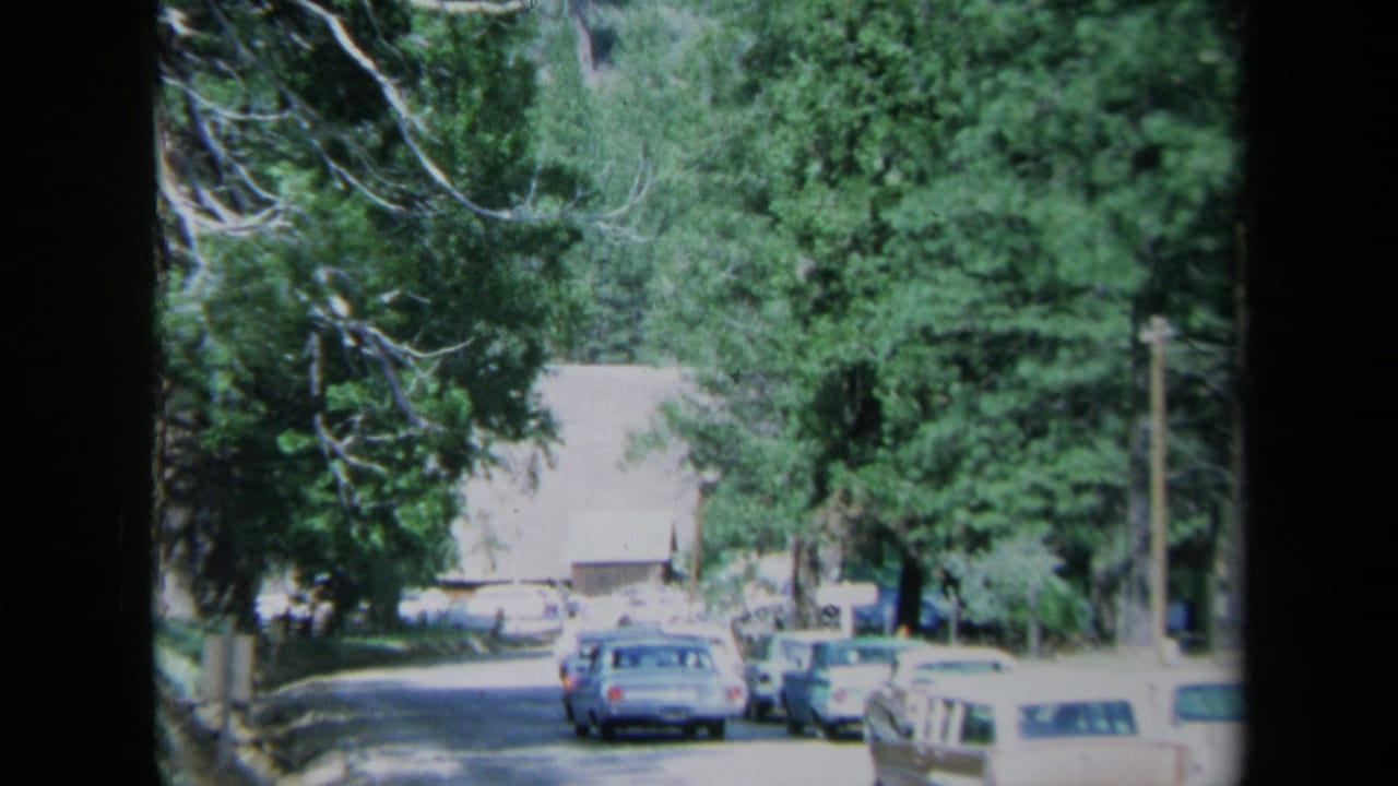 1969 California home-movie frame showing a sunlit tree-lined town street, parked cars, and a rustic wooden building beyond the road.