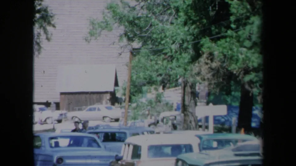 1969 California home-movie still with pedestrians near parked cars on a small-town street, with trees and a rustic building behind them.