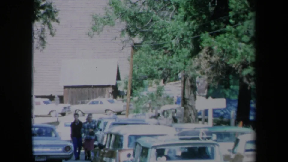1969 California frame showing two pedestrians walking beside parked cars on a tree-lined town street with a wooden building behind them.