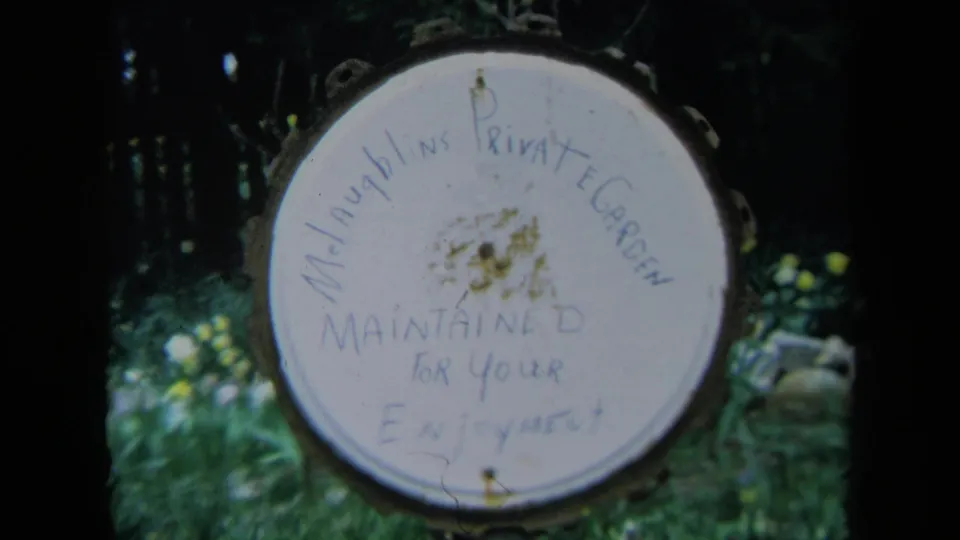 1969 California 8mm frame showing a round hand-painted McLaughlin's Private Garden sign surrounded by shaded greenery.