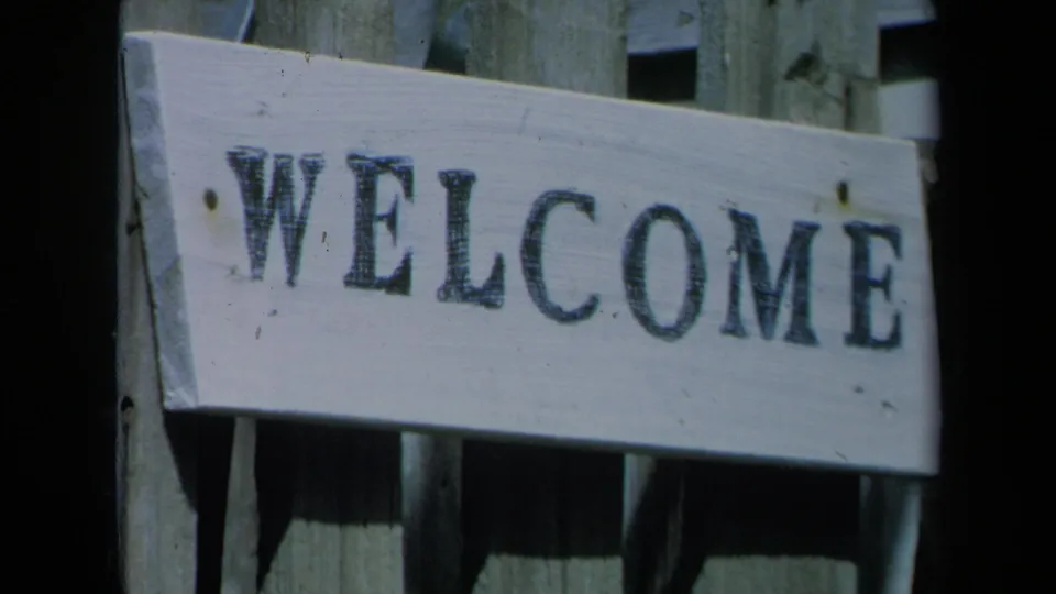 Weathered wooden fence with a pale board reading WELCOME, filmed in a 1969 California home-movie garden scene.