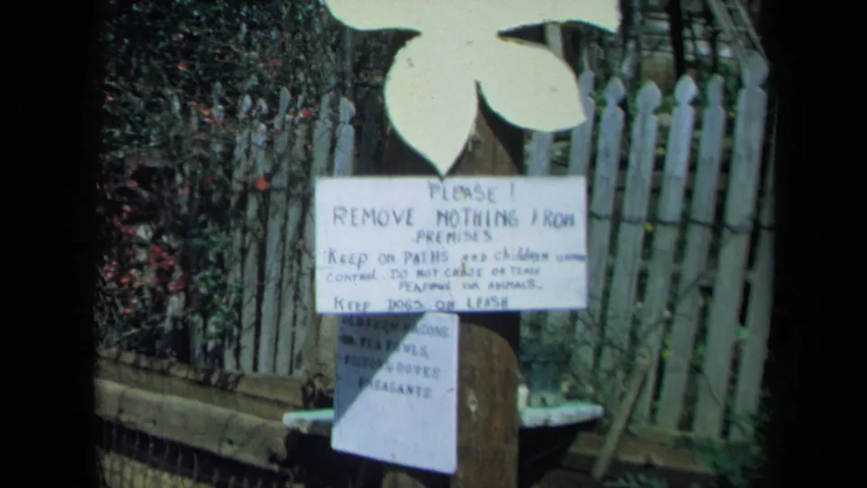 1969 California home-movie frame of a posted garden rule board with a smaller animal list below it near a picket fence.