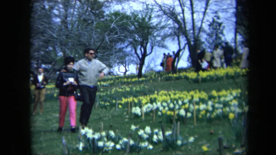 The man and woman continue across the California garden scene, framed by yellow and white flowers, trees, and distant park visitors.
