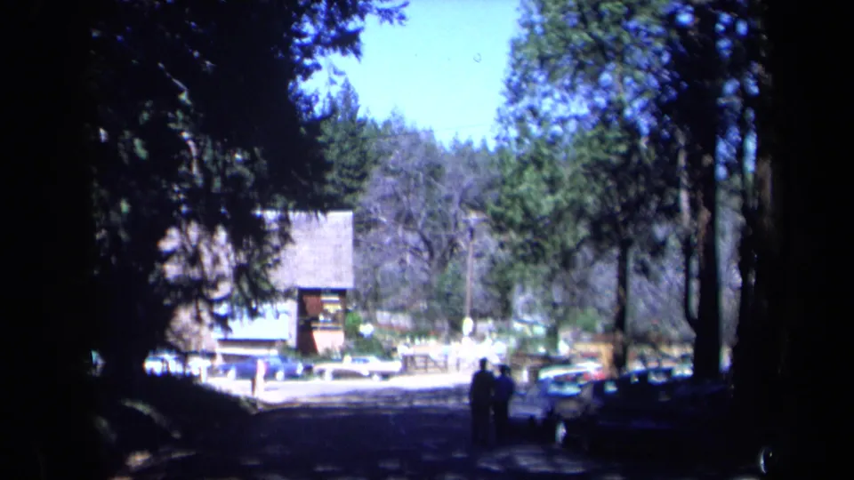 Two distant people walk along a shaded California park path in 1969 footage, with parked cars, tall trees, and a rustic building ahead.