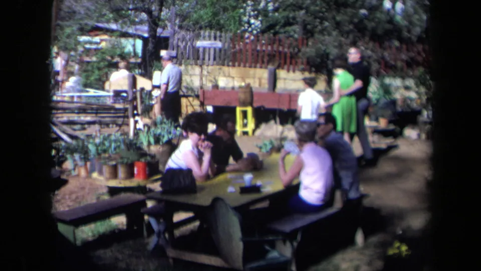 Adults sit and stand around picnic tables in a California garden setting during a 1969 home-movie outing.