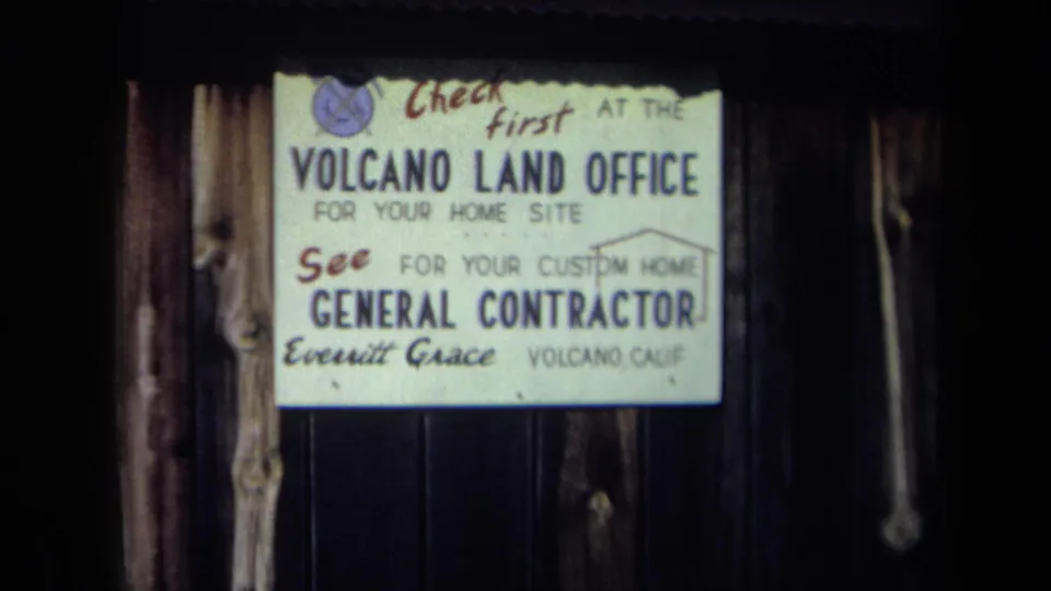 1969 California 8mm frame with a readable Volcano Land Office sign and contractor details on a dark timber wall.