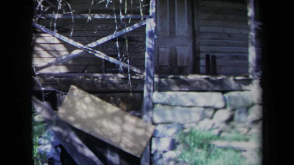 1969 California 8mm frame showing the cabin wall, porch supports, crossed rails, stone foundation, doorway, vines, and overgrown grass.