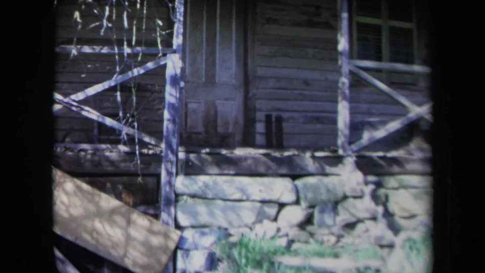 1969 California 8mm frame showing a weathered cabin exterior with a door, window, crossed rail, stone foundation, vines, and grass.