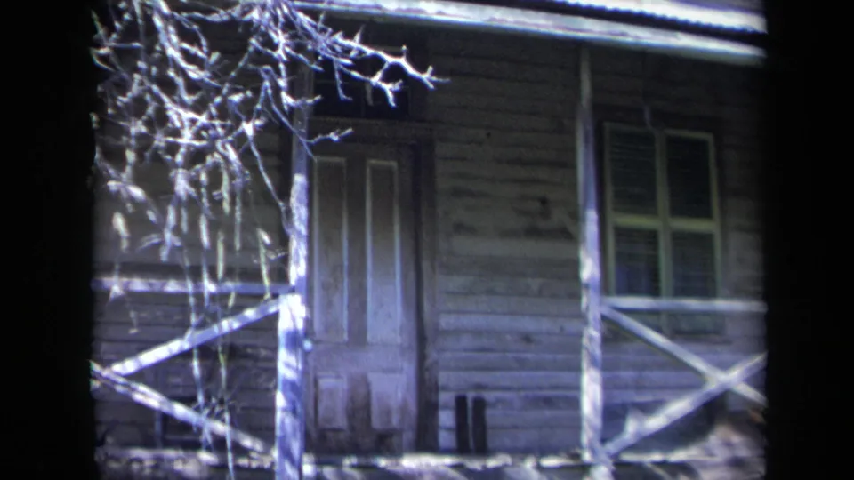 Blue-tinted 1969 California home-movie frame of a closed door, nearby window, porch post, and cross-braced railing on an old wooden house.