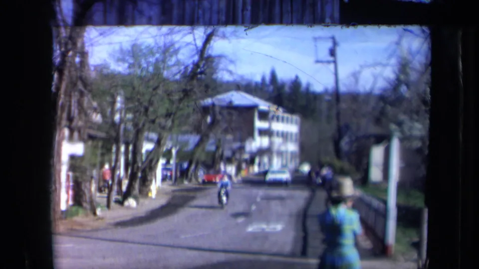1969 California 8mm street view with a cyclist on the road, parked cars, pedestrians, trees, and a multi-story building ahead.