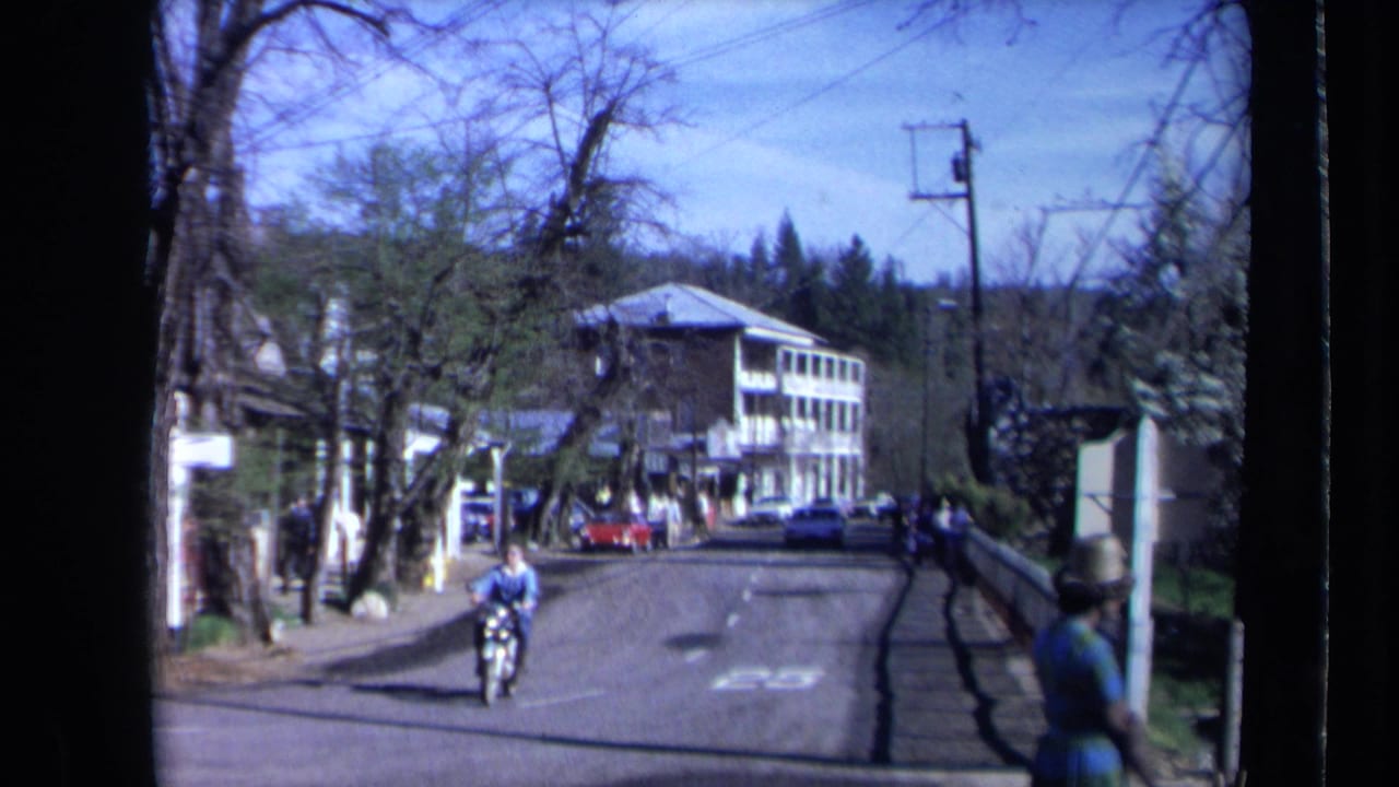 1969 California home-movie frame showing a rider on a small motorbike approaching along a paved road lined with trees, sidewalks, parked cars, and shops.