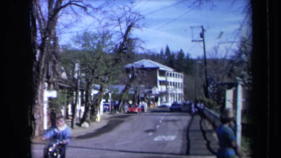 1969 California street frame with a motorbike rider near the foreground and pedestrians, parked cars, trees, and a multi-story building farther down the road.