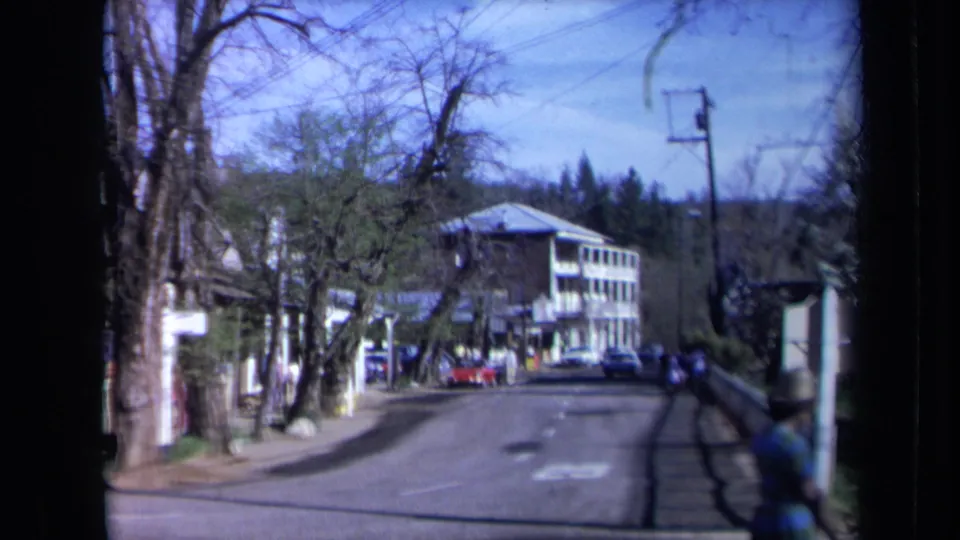1969 California home-movie view of a quiet paved road after the rider has passed, showing trees, sidewalks, parked cars, and a commercial-looking building.