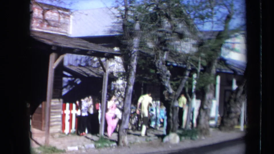 1969 California street-side frame showing pedestrians gathered near a shopfront or outdoor display, with clothing racks, a large tree, and roadside structures.