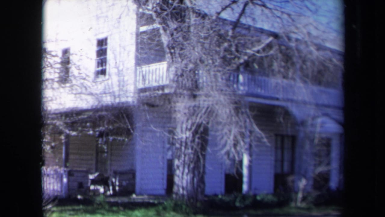 Two-story wooden house with porch columns, balcony, trees, lawn, and dark film borders.