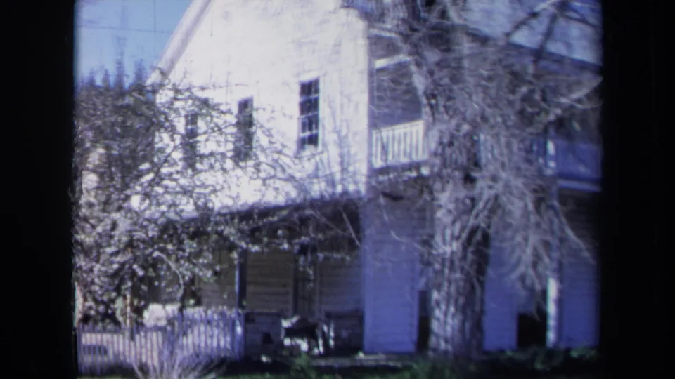 1969 California frame of a wooden house exterior with a white picket fence, balcony, porch area, windows, and bare branches.