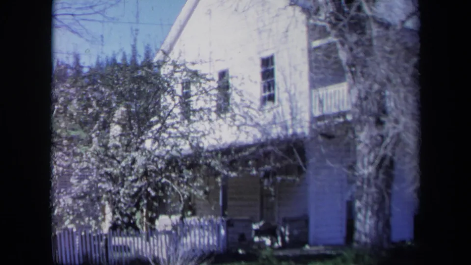 1969 California home-movie frame showing the house facade with picket fence, balcony, porch columns, windows, and heavy tree branches.