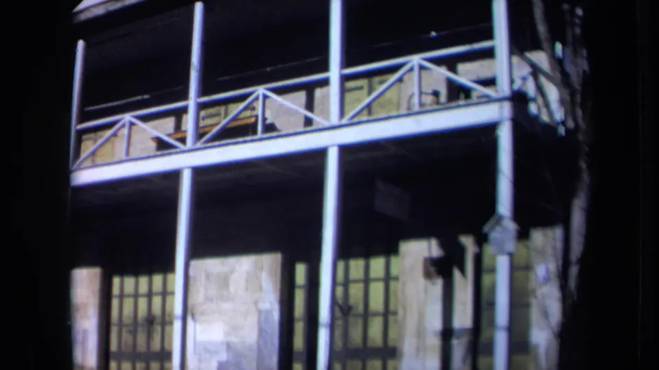 1969 California frame with the camera still on the hall exterior, showing the balcony rail, posts, stone wall, and shaded lower openings.