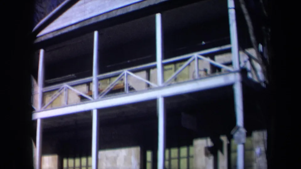 1969 California 8mm frame looking up at the hall's roofline, balcony structure, white posts, and shaded stone facade.