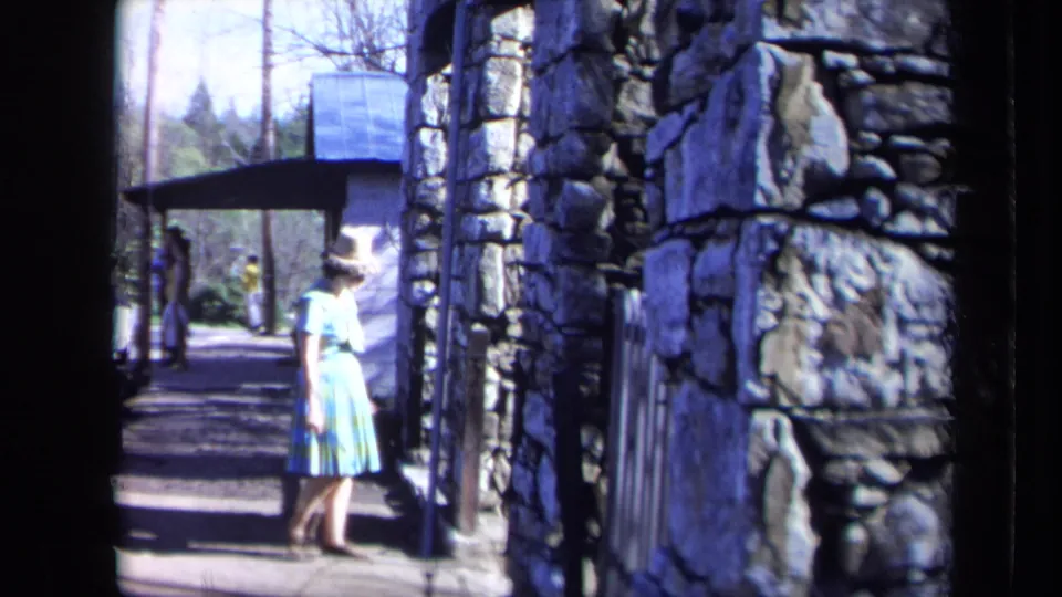 1969 California home-movie frame of a woman in a striped dress and hat beside a rustic stone wall and shaded walkway.