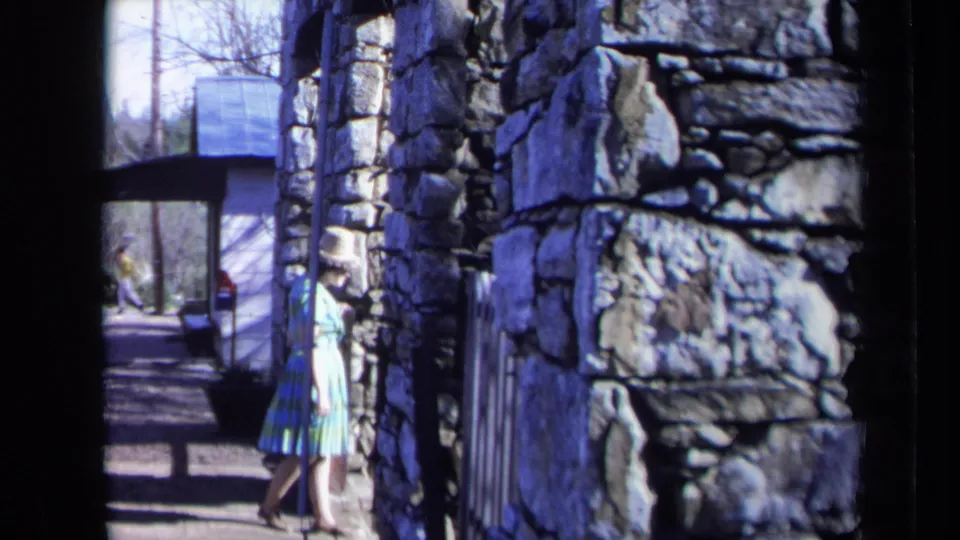 1969 California 8mm frame showing a woman in a hat walking beside rustic masonry and a shaded garden path.
