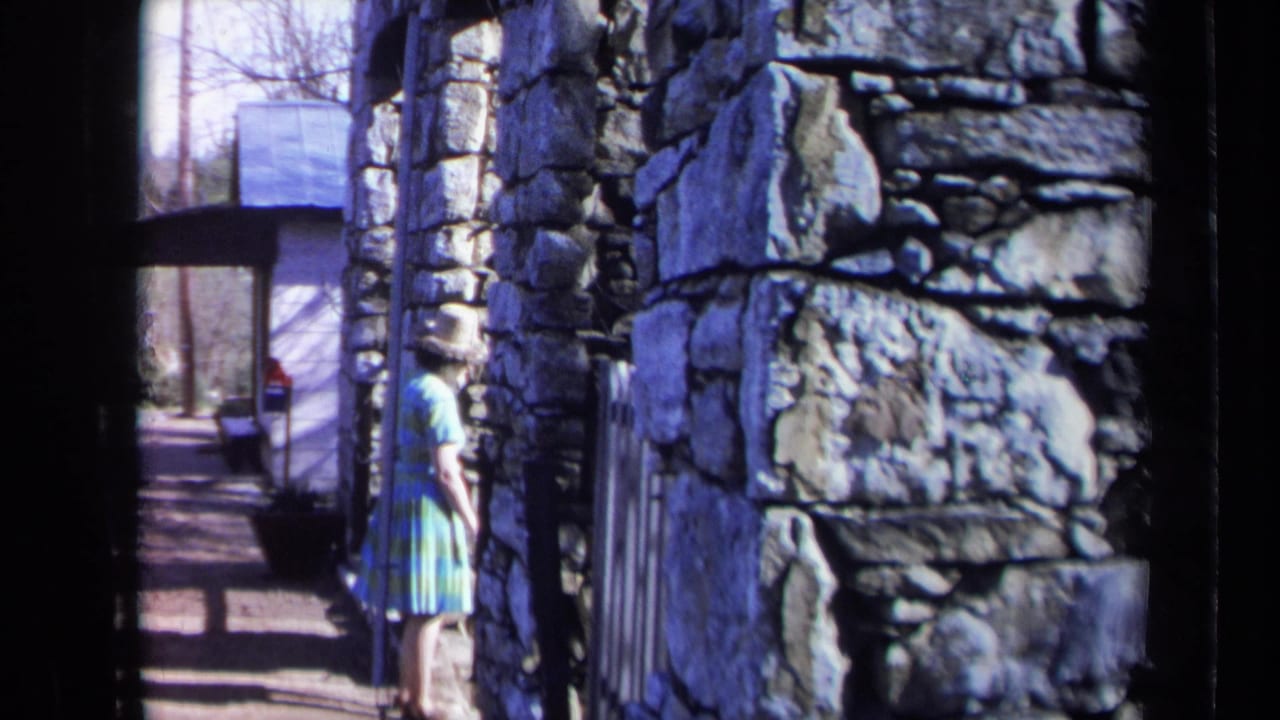1969 California home-movie frame showing a woman walking along a sidewalk beside a rough stone wall and nearby parked car.