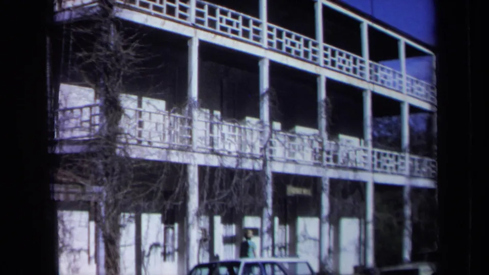 1969 California home-movie view of a sunlit inn facade with white columns, decorative balconies, vines, and a car at ground level.