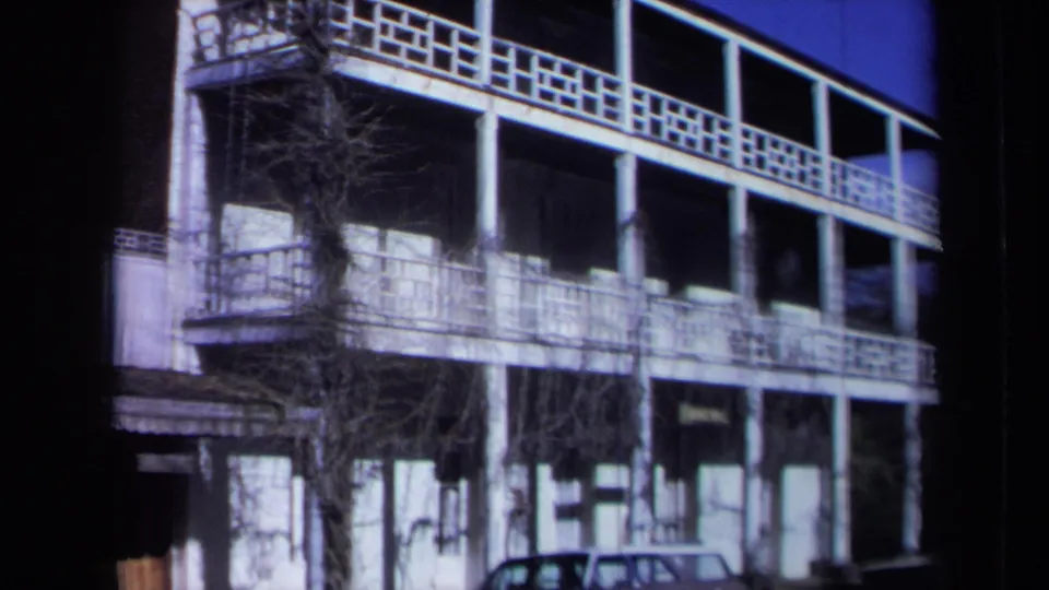 1969 California inn exterior seen from across the street, showing white railings, columns, vines, and a parked car beneath the balcony.
