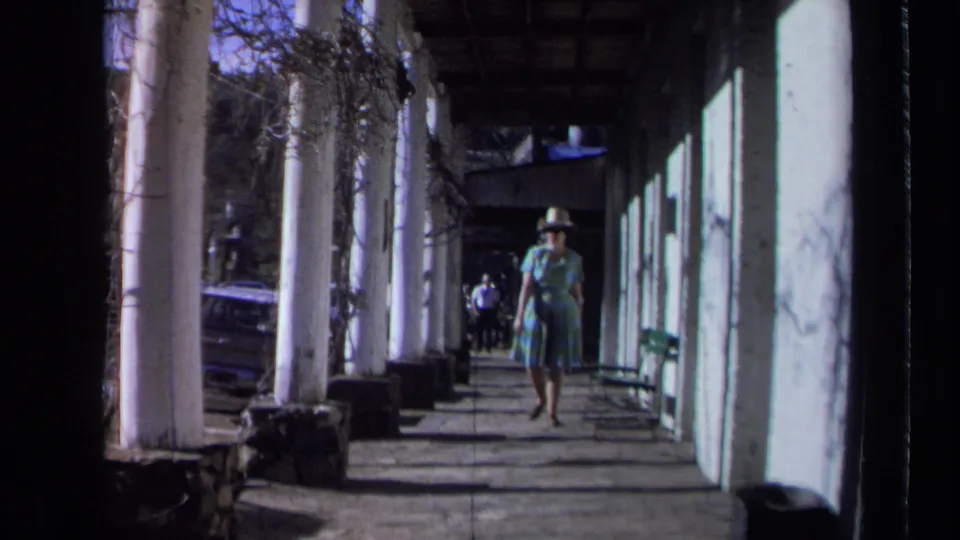 1969 California home-movie frame of a woman in a wide-brimmed hat and dress walking toward the camera along a sunlit covered walkway.