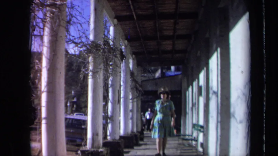 A woman in a hat and patterned dress approaches the camera beneath the white columns of a California inn walkway in 1969 footage.