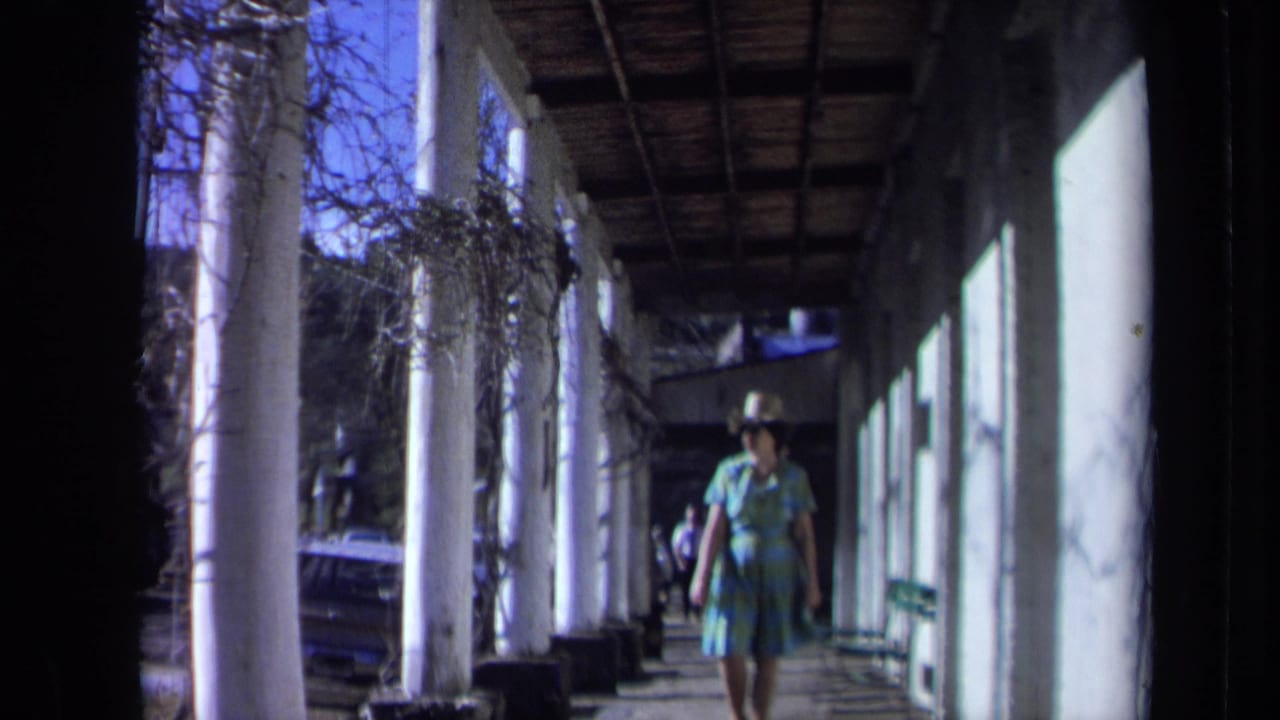 In 1969 California home-movie footage, a woman walks through a shaded colonnade with white columns and a wooden roof.