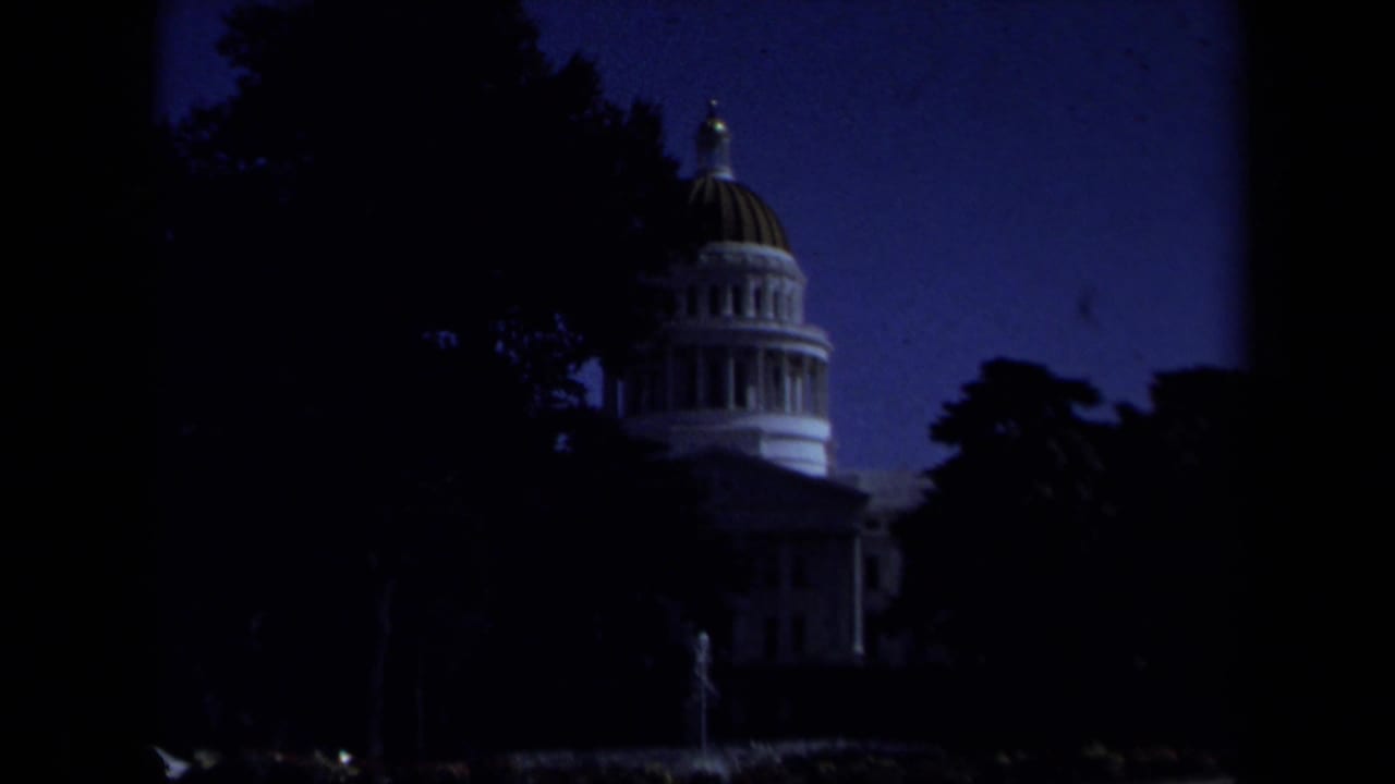 Underexposed 1969 8mm frame of the California State Capitol dome partly obscured by dark trees in Sacramento.
