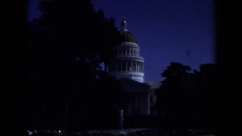 Underexposed 1969 8mm frame of the California State Capitol dome partly obscured by dark trees in Sacramento.