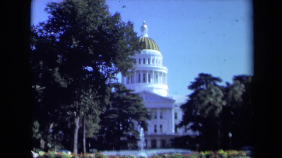 1969 8mm frame showing the California State Capitol dome and white facade partly screened by trees in Sacramento.