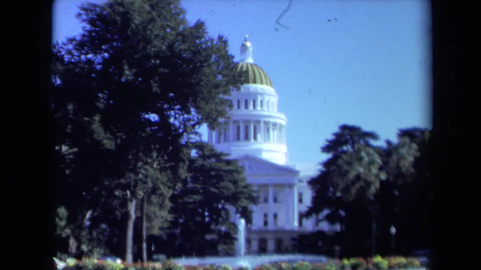 Late-1960s 8mm view of the California State Capitol grounds, with dome, trees, blue sky, and fountain spray visible.