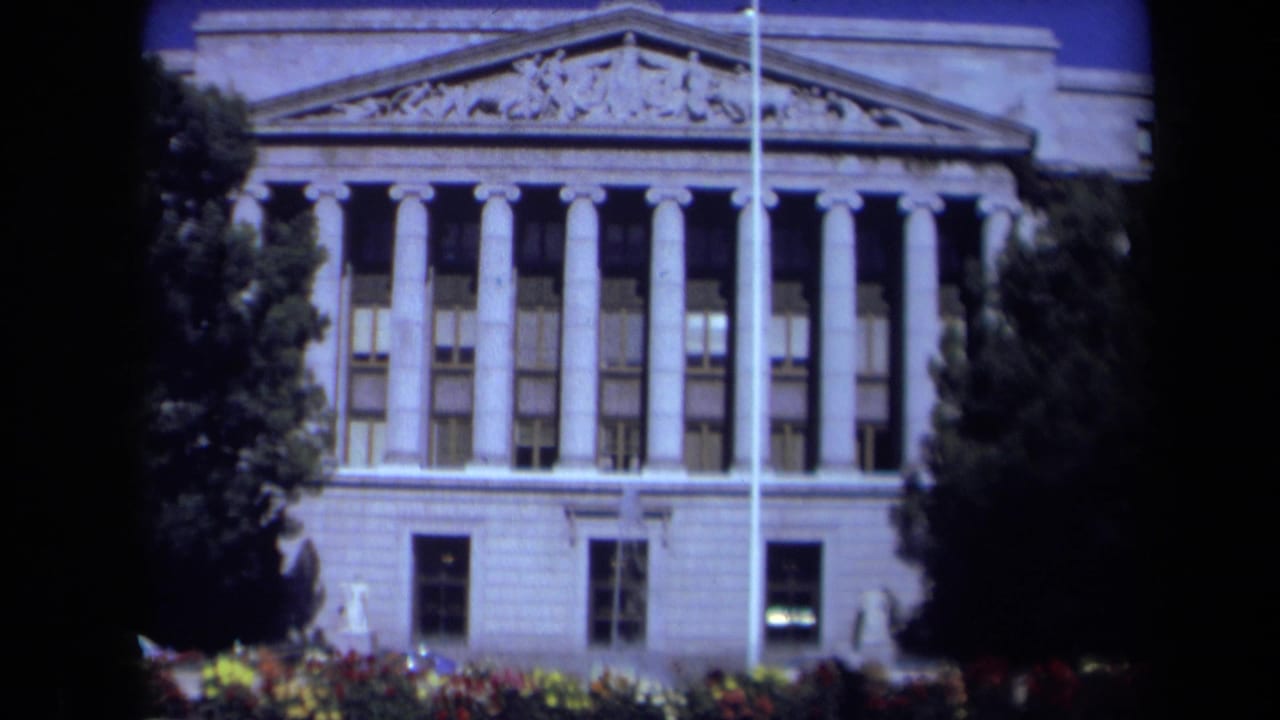 Neoclassical building with tall columns, pediment, trees, and flower beds.