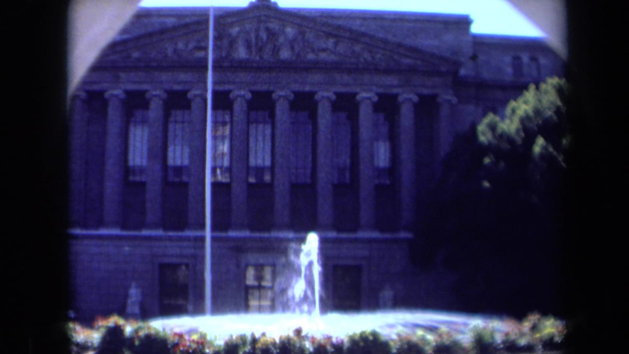 1969 California 8mm frame showing a classical columned facade, flagpole, fountain spray, and landscaped foreground.