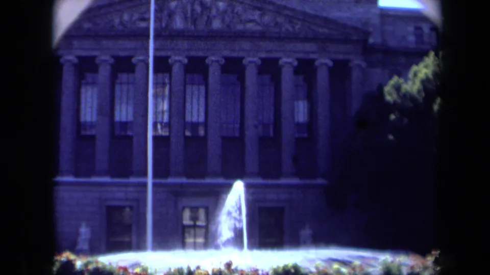 1969 California 8mm frame with a fountain centered before a classical facade, partly framed by tree foliage and film-edge shadows.