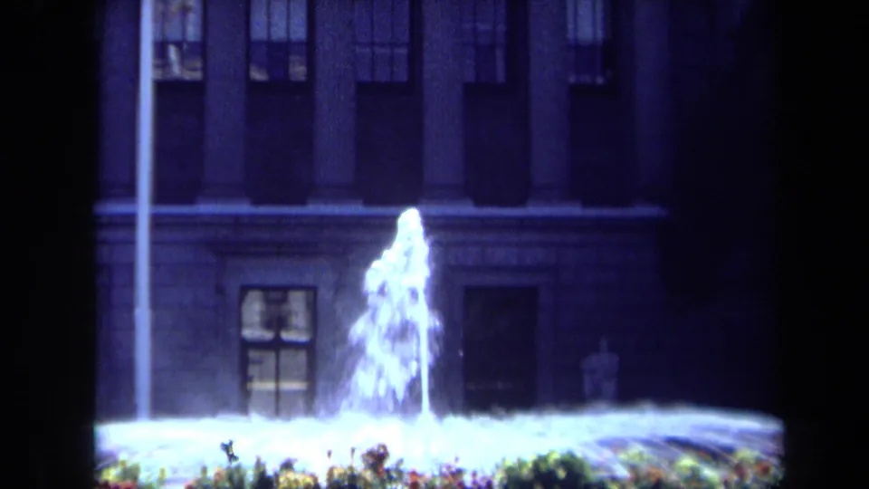 Tight 1969 California 8mm frame of bright fountain spray against the lower columns, windows, and flowerbeds of a classical building.