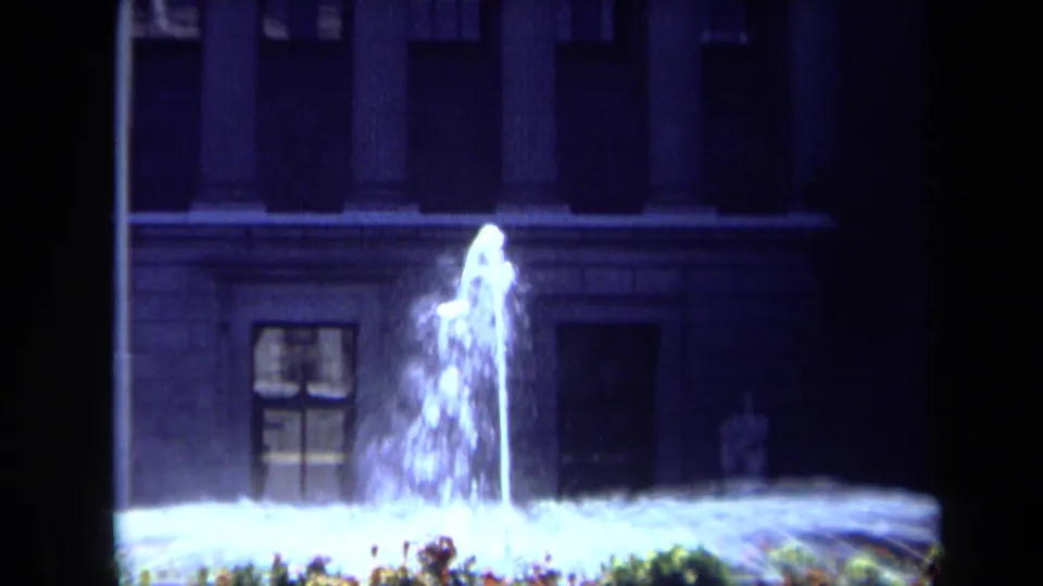 1969 California 8mm frame showing a lit fountain jet, flower bed, and shadowed classical-style building at night.