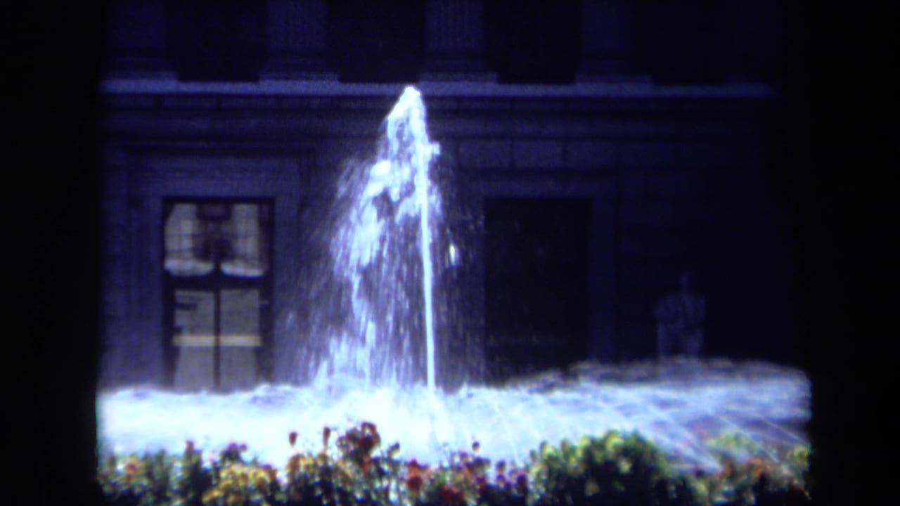 1969 California home-movie frame with a tall illuminated fountain plume, low flower bed, and shadowed building exterior.