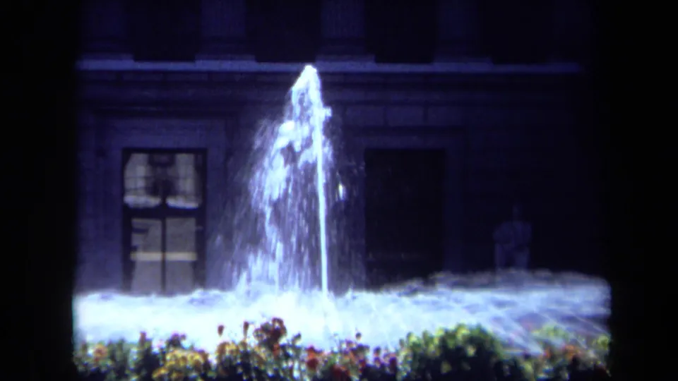 1969 California home-movie frame with a tall illuminated fountain plume, low flower bed, and shadowed building exterior.