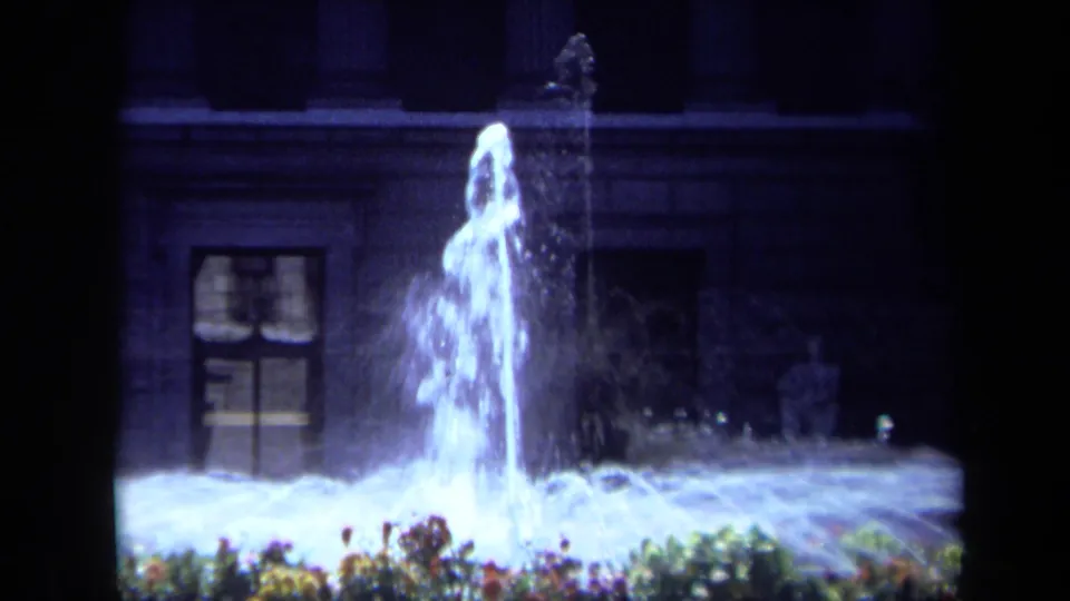Low-light 1969 California 8mm frame of an illuminated fountain display, flower border, and dark architectural backdrop.