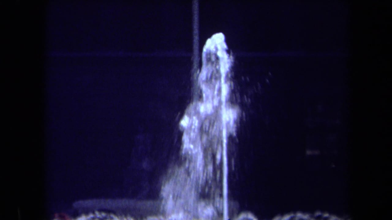 Closer nighttime view of the fountain's top spout with water falling around it.