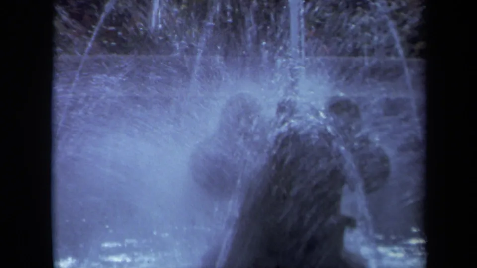 1969 California 8mm frame of an illuminated fountain with arcing jets, mist, and a dark background.